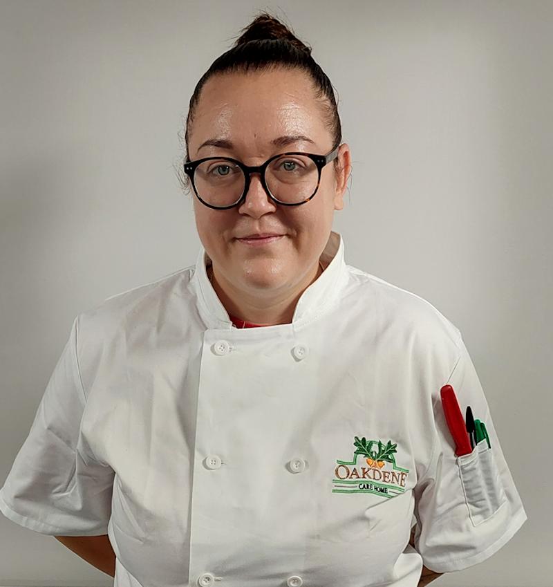 female head chef with brown hair in a white uniform smiling for her team photo