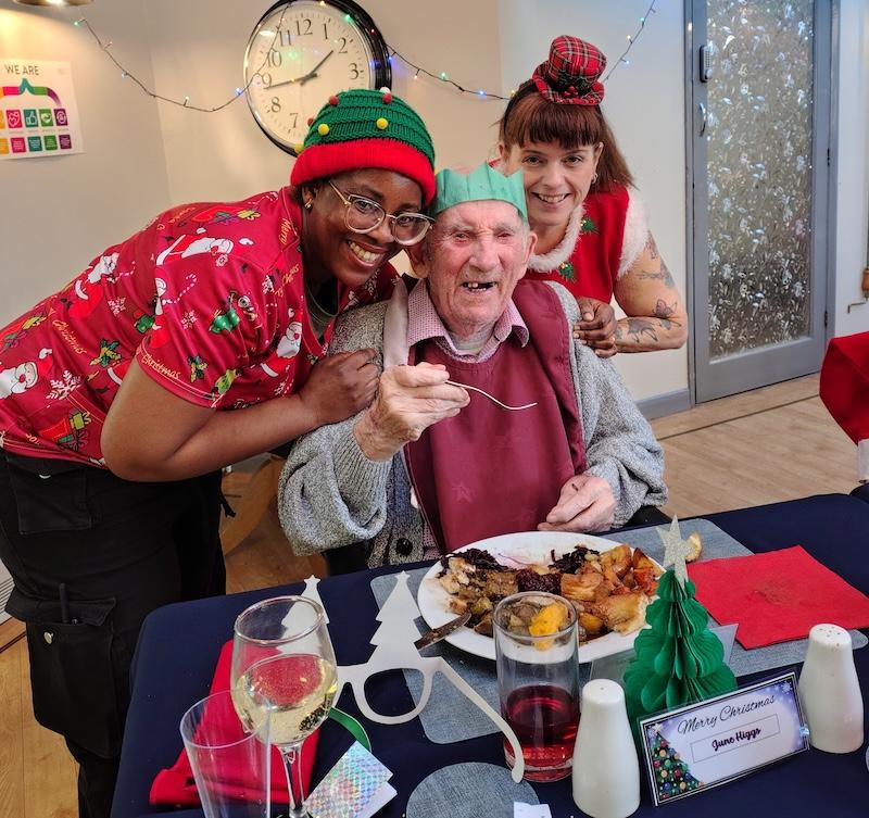 2 female care workers dressed in Christmas attire stood next to an elderly man enjoying Christmas dinner