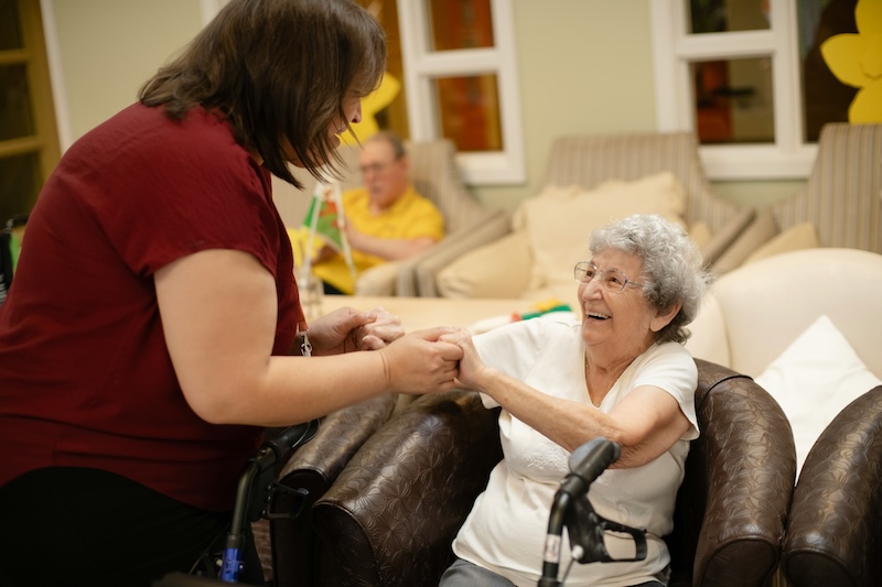 female resident sat down in an armchair holding hands and smiling with a carer